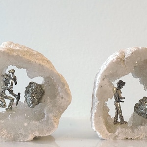 May include: Two white geode sculptures, each with a hollowed-out center. One geode features a silver figurine of a person running, the other a person standing. Both geodes have a small, round, silver-colored stone.