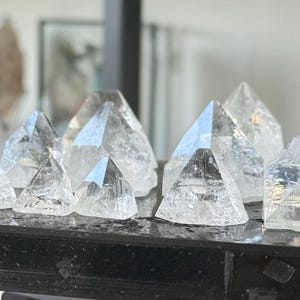 May include: A collection of clear, faceted quartz crystals shaped into small pyramids. Each crystal features a pointed apex and multiple triangular faces, displaying natural internal inclusions and transparency, arranged on a dark surface.