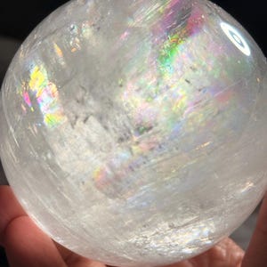 May include: A spherical clear quartz crystal ball featuring internal fractures and inclusions that create vibrant rainbow light refractions across its surface. The polished stone has a smooth, rounded shape with visible natural patterns inside.