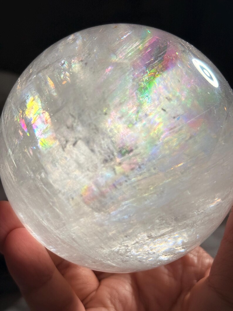 May include: A spherical clear quartz crystal ball featuring internal fractures and inclusions that create vibrant rainbow light refractions across its surface. The polished stone has a smooth, rounded shape with visible natural patterns inside.