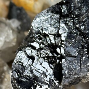 May include: Close-up of a dark, reflective mineral specimen with a crystalline structure. The surface is a deep black with areas of high reflectivity, creating a shimmering effect. The mineral appears to be a rough, natural stone.