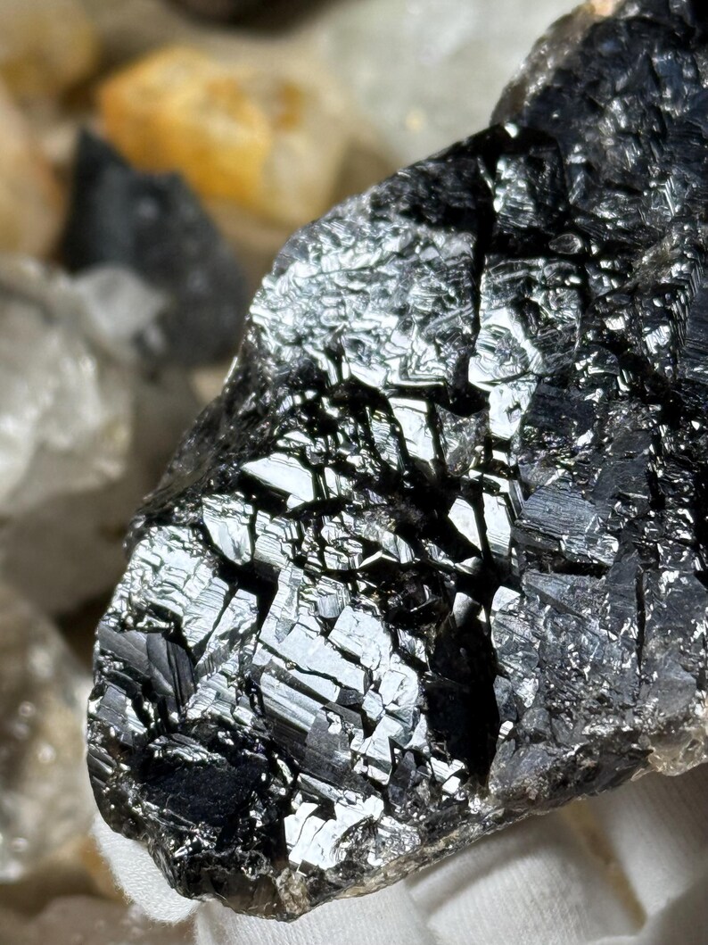 May include: Close-up of a dark, reflective mineral specimen with a crystalline structure. The surface is a deep black with areas of high reflectivity, creating a shimmering effect. The mineral appears to be a rough, natural stone.