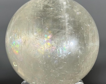 May include: A polished, spherical clear quartz crystal ball featuring internal fractures and inclusions that create vibrant, multi-colored rainbow light refractions across its translucent, pale off-white surface.