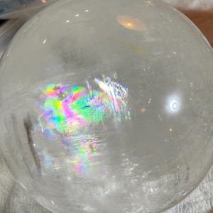 May include: A clear, spherical quartz crystal ball featuring internal fractures that create vibrant, multi-colored rainbow light refractions. The surface is smooth and polished, reflecting ambient light with bright highlights across its rounded form.