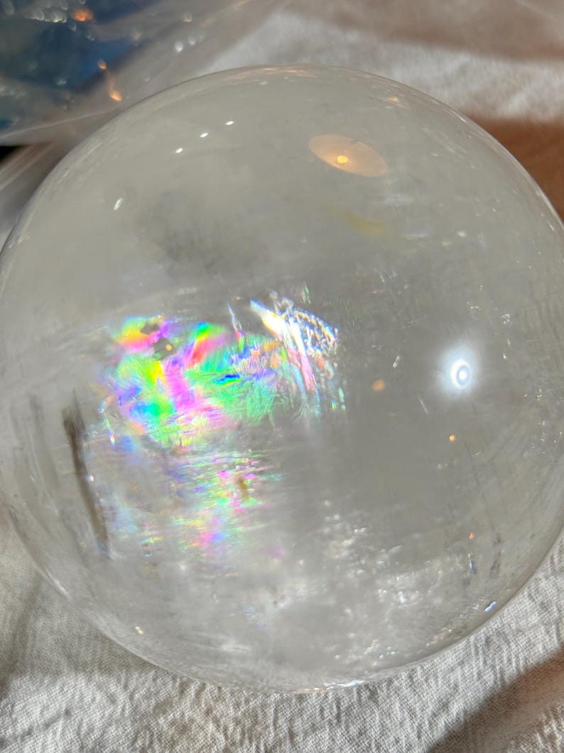 May include: A clear, spherical quartz crystal ball featuring internal fractures that create vibrant, multi-colored rainbow light refractions. The surface is smooth and polished, reflecting ambient light with bright highlights across its rounded form.