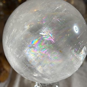 May include: A clear, spherical quartz crystal ball featuring internal fractures and inclusions that create a vibrant, multi-colored rainbow refraction effect across its polished, smooth, and translucent surface under bright lighting.