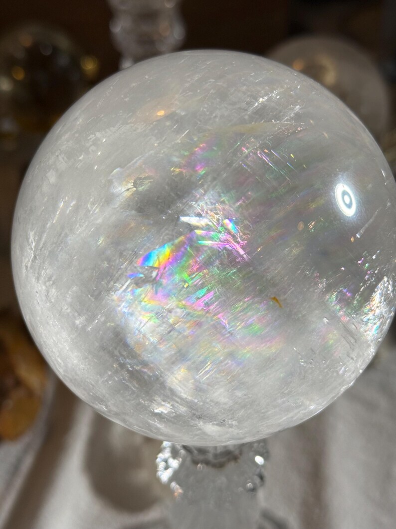 May include: A clear, spherical quartz crystal ball featuring internal fractures and inclusions that create a vibrant, multi-colored rainbow refraction effect across its polished, smooth, and translucent surface under bright lighting.
