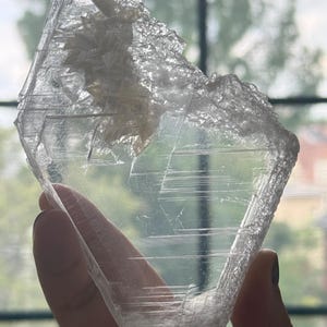 May include: A translucent, clear selenite crystal specimen with a flat, blade-like shape. The mineral features visible internal striations, linear etchings, and a textured, jagged crystalline formation along the upper edge of the structure.