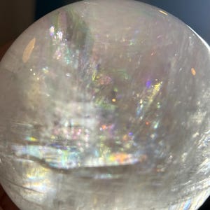 May include: A clear, spherical crystal ball featuring internal fractures and inclusions that create a multi-colored, iridescent rainbow effect when light passes through the translucent, polished surface of the stone sphere.