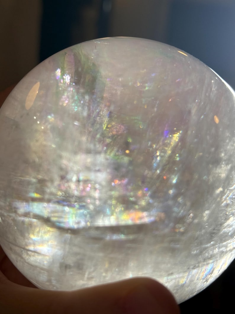 May include: A clear, spherical crystal ball featuring internal fractures and inclusions that create a multi-colored, iridescent rainbow effect when light passes through the translucent, polished surface of the stone sphere.