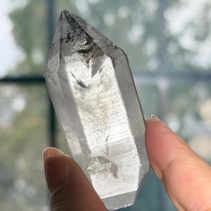 May include: A clear, hexagonal quartz crystal point featuring a sharp, terminated tip and internal inclusions. The translucent stone displays natural facets and a smooth, polished surface, with dark mineral deposits visible near the apex.
