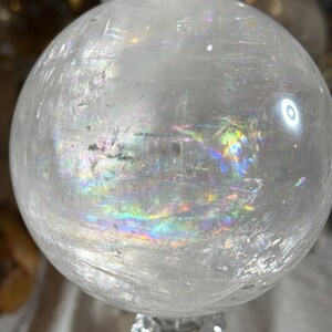 May include: A clear, spherical quartz crystal ball featuring internal fractures and inclusions that create vibrant, multi-colored rainbow light refractions throughout the polished, translucent surface of the stone.