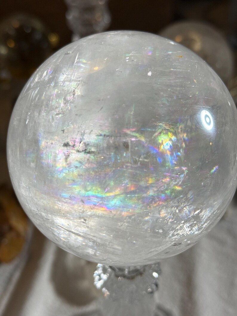 May include: A clear, spherical quartz crystal ball featuring internal fractures and inclusions that create vibrant, multi-colored rainbow light refractions throughout the polished, translucent surface of the stone.