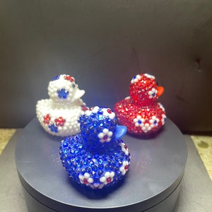 Op de afbeelding: Drie decoratieve rubberen eendjes. E&eacute;n is blauw, &eacute;&eacute;n is rood en &eacute;&eacute;n is wit. Elke eend is bedekt met glinsterende strass-steentjes en versierd met kleine bloemaccenten in contrasterende kleuren. De eendjes staan op een donkergrijze ondergrond.