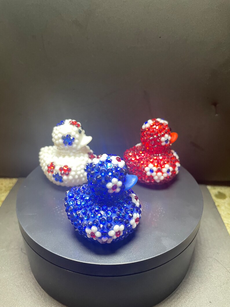 Op de afbeelding: Drie decoratieve rubberen eendjes. E&eacute;n is blauw, &eacute;&eacute;n is rood en &eacute;&eacute;n is wit. Elke eend is bedekt met glinsterende strass-steentjes en versierd met kleine bloemaccenten in contrasterende kleuren. De eendjes staan op een donkergrijze ondergrond.