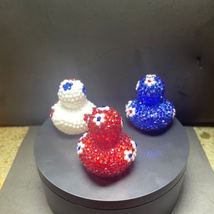 Op de afbeelding: Drie decoratieve eendenbeeldjes, elk bedekt met glinsterende kralen. E&eacute;n is rood, een andere wit en de derde blauw. Elke eend heeft kleine witte en blauwe bloemaccenten. De eenden worden gepresenteerd op een donkergrijs draaiend platform.