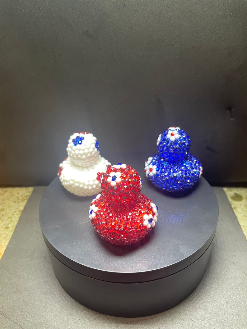 Op de afbeelding: Drie decoratieve eendenbeeldjes, elk bedekt met glinsterende kralen. E&eacute;n is rood, een andere wit en de derde blauw. Elke eend heeft kleine witte en blauwe bloemaccenten. De eenden worden gepresenteerd op een donkergrijs draaiend platform.