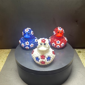 Op de afbeelding: Drie decoratieve badeenden, elk versierd met glinsterende versieringen. E&eacute;n is blauw, &eacute;&eacute;n is rood en &eacute;&eacute;n is wit, allemaal met een bloemmotief in rood, wit en blauw. De eenden worden gepresenteerd op een donkergrijs draaiend platform.