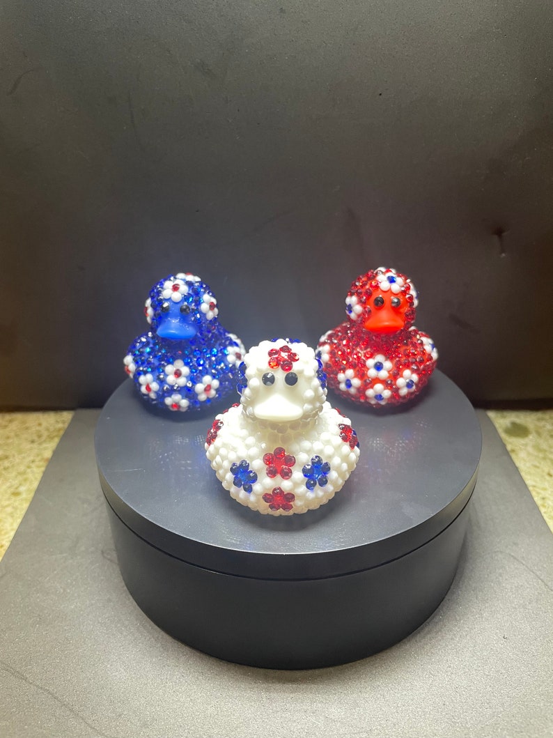 Op de afbeelding: Drie decoratieve badeenden, elk versierd met glinsterende versieringen. E&eacute;n is blauw, &eacute;&eacute;n is rood en &eacute;&eacute;n is wit, allemaal met een bloemmotief in rood, wit en blauw. De eenden worden gepresenteerd op een donkergrijs draaiend platform.