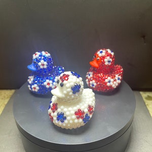 Op de afbeelding: Drie decoratieve badeenden. E&eacute;n is wit met rode en blauwe bloemaccenten, een andere is blauw met witte en rode bloemaccenten en de derde is rood met witte en blauwe bloemaccenten. Elke eend heeft een snavel van een andere kleur.