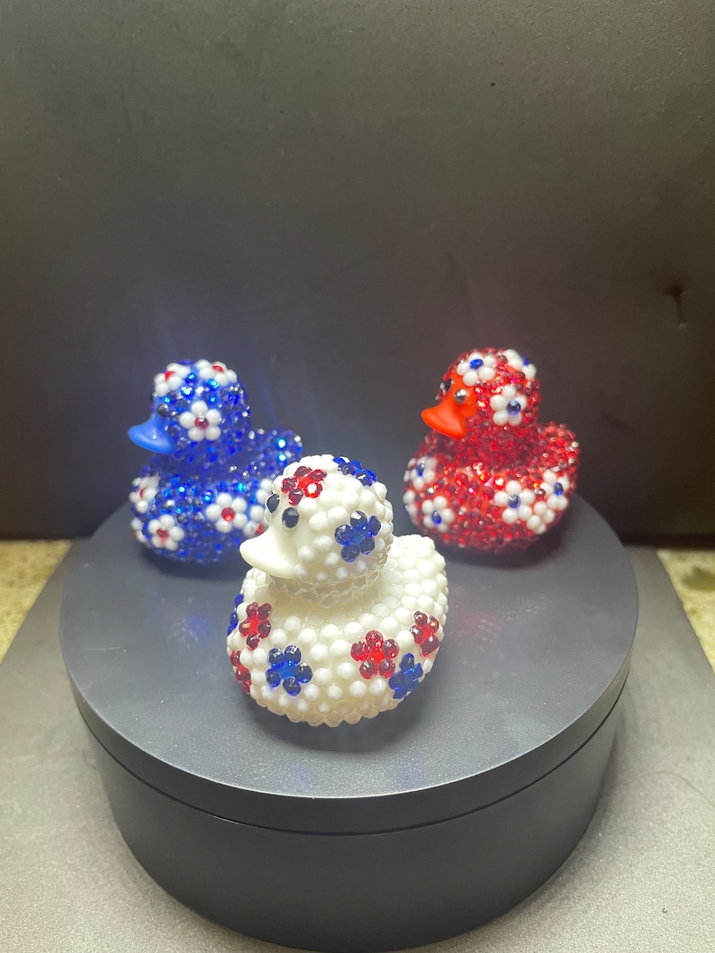 Op de afbeelding: Drie decoratieve badeenden. E&eacute;n is wit met rode en blauwe bloemaccenten, een andere is blauw met witte en rode bloemaccenten en de derde is rood met witte en blauwe bloemaccenten. Elke eend heeft een snavel van een andere kleur.