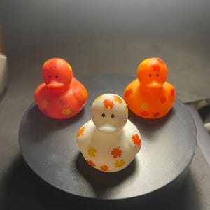 May include: Three rubber duck toys with fall leaf designs. One duck is orange with orange and yellow leaves, one is red with orange and yellow leaves, and one is white with orange and yellow leaves.