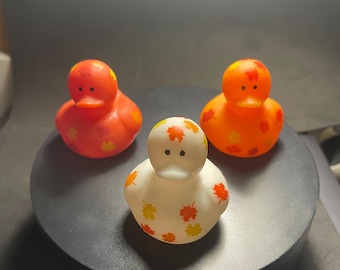May include: Three rubber duck toys with fall leaf designs. One duck is orange with orange and yellow leaves, one is red with orange and yellow leaves, and one is white with orange and yellow leaves.