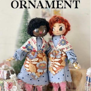 Op de afbeelding: Twee folk doll ornamenten. De poppen hebben zwart en rood haar, dragen blauwe jurken met bloemen schorten en sjaals. De tekst "FOLK DOLL ORNAMENT" staat bovenaan de afbeelding. De poppen staan in de buurt van ingepakte cadeaus.