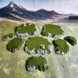 Puede incluir: Un conjunto de siete piezas de terreno en miniatura de color verde y gris para juegos de mesa. Las piezas tienen forma de colinas rocosas con cimas cubiertas de hierba.