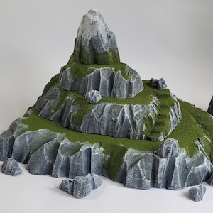 Puede incluir: Un terreno de montaña modelo gris y verde con un pico alto y puntiagudo. El terreno está hecho de espuma texturizada y está diseñado para juegos de mesa o juegos de guerra en miniatura.