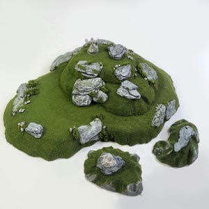 Puede incluir: Un conjunto de terreno en miniatura verde y gris para juegos de mesa. El conjunto incluye una gran colina cubierta de hierba con varias rocas grises y una formación rocosa más pequeña y separada. La colina es perfecta para usar en juegos de fantasía o medievales.
