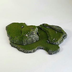 Puede incluir: Una pieza de terreno en miniatura verde y gris con una superficie rocosa y cubierta de hierba. La pieza está diseñada para juegos de mesa y presenta una pequeña colina y un camino sinuoso.