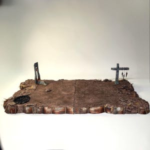 May include: A diorama depicting a post-apocalyptic landscape. The scene features a weathered, brown terrain with a guillotine and a cross with hanging figures. The base includes corrugated metal structures, suggesting a war-torn environment. The overall color palette is muted, with shades of brown and gray.