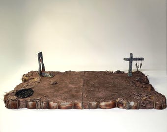 Pode incluir: Diorama retratando uma paisagem pós-apocalíptica. A cena apresenta um terreno castanho e desgastado com uma guilhotina e uma cruz com figuras penduradas. A base inclui estruturas de metal ondulado, sugerindo um ambiente devastado pela guerra. A paleta de cores é suave, com tons de castanho e cinza.
