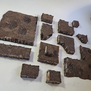 Może przedstawiać: Zestaw 12 brązowych i beżowych miniaturowych elementów terenu do gier planszowych. Elementy wykonane są z tektury i mają fakturowaną powierzchnię przypominającą ziemię i drewno. Elementy są zaprojektowane tak, aby reprezentować okopy, bunkry i inne fortyfikacje.