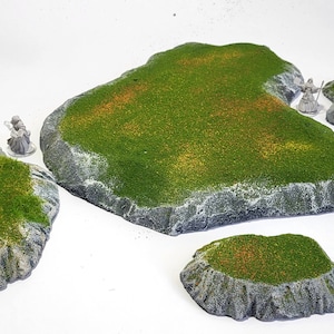 Puede incluir: Cinco islas miniatura verdes y grises con bordes rocosos, perfectas para juegos de mesa. Las islas están cubiertas de hierba verde con parches de marrón y amarillo.
