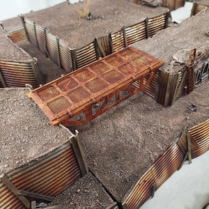 May include: A miniature, weathered, rusty-brown metal bridge spans a gap between two earthen trenches. The bridge has a grid-like surface and is supported by a triangular frame. The trenches are lined with corrugated metal and wooden supports, creating a war-torn scene.