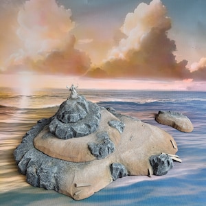 Puede incluir: Una figura en miniatura de una persona de pie en una isla rocosa, con una playa de arena y un océano azul en el fondo.