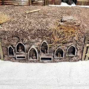 May include: A miniature, rustic building with a thatched roof and wooden accents. The structure features arched openings along the front and is decorated with dried grass. The building is likely a model for a diorama or tabletop game.