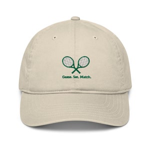 May include: A beige baseball cap with green embroidered tennis rackets and the text "Game. Set. Match."