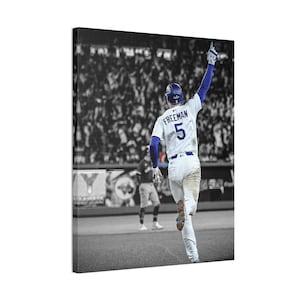 以下が含まれることがあります： 白と青のユニフォームを着た野球選手、背番号5、背中に「FREEMAN」と書かれたキャンバスプリント。選手は腕を上げてお祝いしています。背景は白黒のスタジアムのシーンです。
