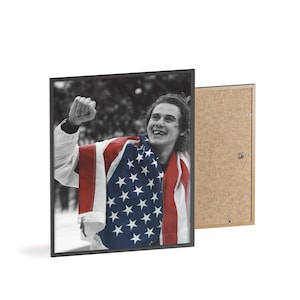 May include: Framed black and white photograph of a person wrapped in an American flag, celebrating. The photograph is in a black frame, with a visible backboard. The person is smiling and has their fist raised in the air.