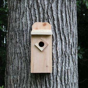 May include: A wooden birdhouse with a natural finish is mounted on a tree trunk. The birdhouse features a diamond-shaped entrance hole and a small roof. The birdhouse is made of light-colored wood.