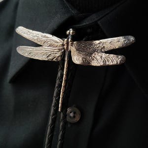 Könnte beinhalten: Eine silberfarbene Bolo-Krawatte mit einer Libelle. Die Libelle hat detaillierte Flügel und einen Körper, die an einem schwarzen geflochtenen Lederband befestigt sind. Die Krawatte ist an einem dunkelgrünen Button-Down-Hemd befestigt.