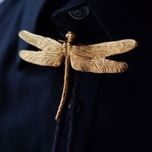 May include: A gold-coloured dragonfly bolo tie. The dragonfly has detailed, textured wings and body. The tie is attached to a black shirt.