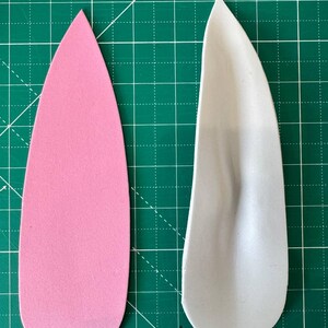 May include: Two foam ear shapes, one pink and one gray, are displayed on a green cutting mat. The pink ear is smooth, while the gray one has a molded texture. The shapes are for a craft project.