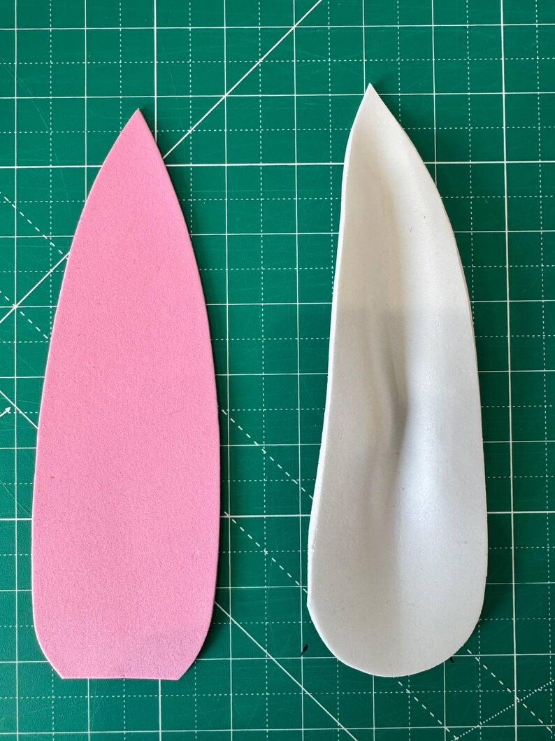 May include: Two foam ear shapes, one pink and one gray, are displayed on a green cutting mat. The pink ear is smooth, while the gray one has a molded texture. The shapes are for a craft project.