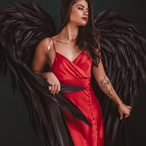 May include: A red satin dress with a high slit and button details. The dress is paired with large black feathered wings. The model has dark hair and red lipstick. A tattoo is visible on the arm.