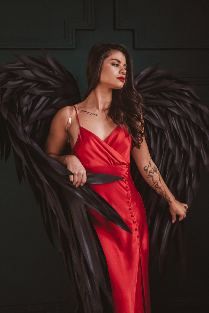May include: A red satin dress with a high slit and button details. The dress is paired with large black feathered wings. The model has dark hair and red lipstick. A tattoo is visible on the arm.