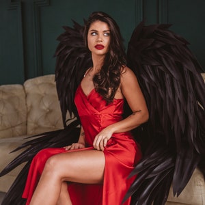 May include: A woman wearing a red satin dress and large black feathered angel wings, seated on a beige sofa. The dress has a high slit, and the wings are spread behind her. The background is a dark green wall.